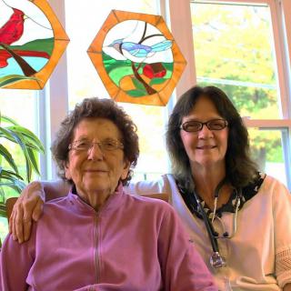 An elderly woman and a younger woman sitting together in front of a window with stained glass art.