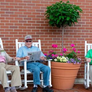 A group of elderly people sitting in rocking chairs outdoors in front of a brick wall, enjoying a relaxed moment with potted flowers nearby.