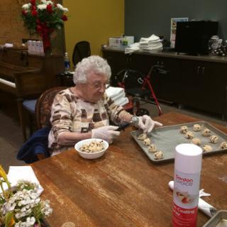An elderly woman carefully prepares cookies at a communal table in a vibrant indoor setting.