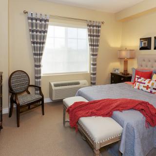 A cozy bedroom with a neatly made bed, accented by red decor, and a window letting in natural light.