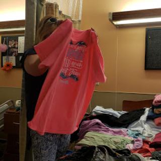 A person holding up a bright pink t-shirt while sorting clothes in a room.