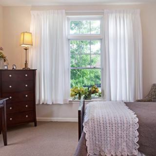 A cozy bedroom with a large window, white curtains, and a wooden dresser.
