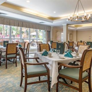 A well-lit dining area with neatly arranged tables and chairs, featuring a large window and chandelier.