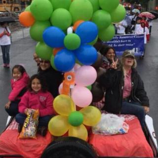 A parade float decorated with colorful balloons carries happy participants on a rainy day.