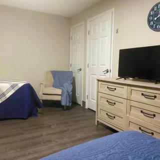 A cozy bedroom featuring a bed with a blue blanket, an armchair, a dresser with a television on top, and wooden flooring.