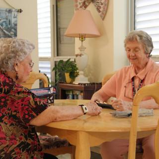 Two elderly women sitting at a table having a conversation in a cozy living room.