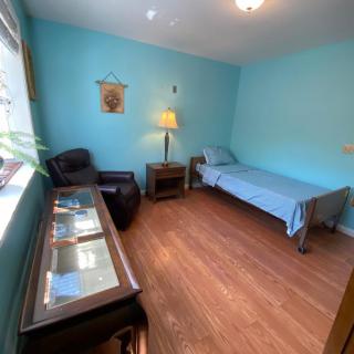 A small bedroom with blue walls features a single bed, armchair, lamp, and a window letting in natural light.