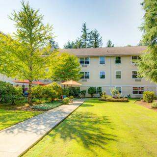 A sunny view of a residential apartment building surrounded by a landscaped garden with trees and a well-maintained lawn.