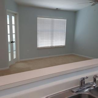 An empty room with a window featuring blinds and a door, viewed from the kitchen area.