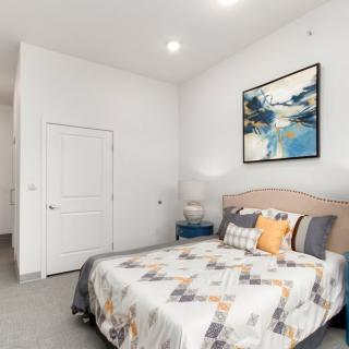 A bright and minimalist bedroom featuring a neatly made bed with patterned bedding, a bedside lamp, and abstract artwork on the wall.