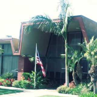 A modern architectural building featuring palm trees and an American flag in a sunny environment.