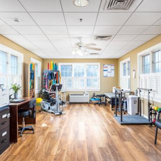 A spacious rehabilitation room filled with therapy and exercise equipment, featuring bright lighting and wooden flooring.