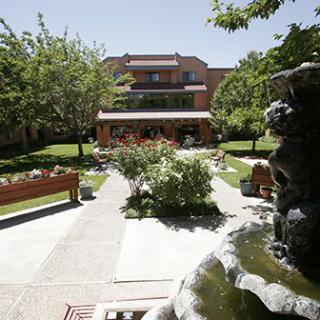 A peaceful garden with a fountain and benches, surrounded by greenery, leading up to a multi-story building.