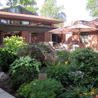 A beautiful garden with lush greenery surrounds a brick house featuring a cozy patio with outdoor furniture and umbrellas.