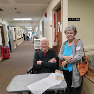 Two elderly individuals smiling together in a care facility hallway.