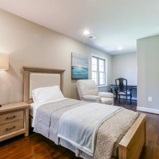 A cozy bedroom with a single bed, nightstand, lamp, armchair, desk, and window.