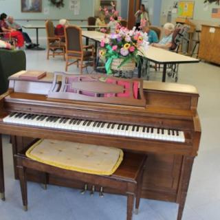 A piano in a communal space with people seated at tables and decorative flowers on display.