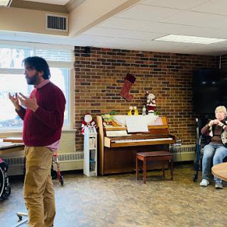 A speaker engages with an audience in a cozy indoor setting, accompanied by a piano.