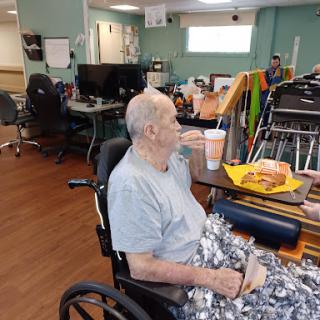 An elderly individual enjoying a snack and beverage in a care facility's activity room.