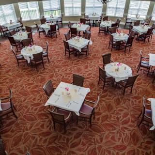 An empty dining area with round tables and chairs, set on a red patterned carpet with large windows in the background.
