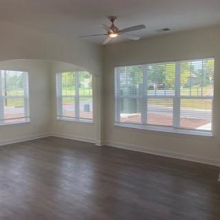 An empty room with large windows and a ceiling fan, featuring hardwood floors and ample natural light.