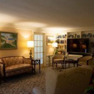 A cozy living room with a sofa, lamps, and a large television, decorated with artwork and books on shelves.