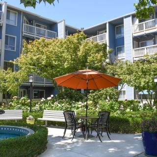 A serene apartment courtyard featuring a patio table with an orange umbrella, surrounded by lush greenery and benches.