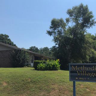 A building with a sign reading 'Methodist Village' surrounded by grass and trees under a clear sky.