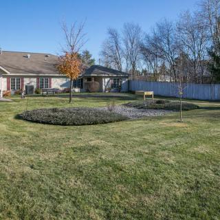 A spacious backyard with a well-maintained lawn, featuring a garden bed and surrounded by trees and a fence under a clear blue sky.