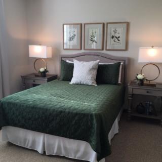 A cozy bedroom featuring a bed with green bedding, flanked by matching side tables and lamps, adorned with elegant wall art.