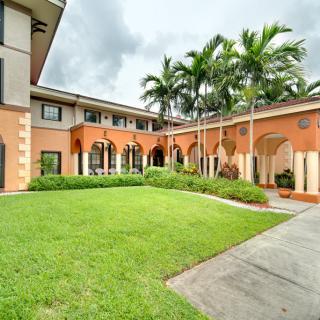A peaceful courtyard surrounded by a building with palm trees and greenery under a cloudy sky.