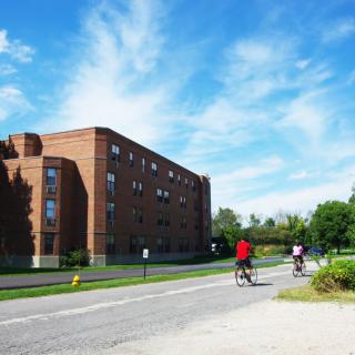 Two cyclists ride past a brick building under a bright blue sky.