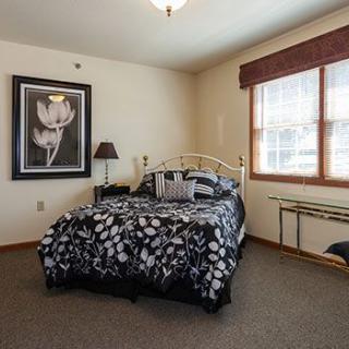 A cozy bedroom with a floral black and white bedspread, a framed artwork on the wall, and natural light streaming through a window.