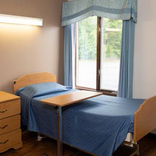 A simple hospital room featuring a bed with blue bedding, a bedside table, and a window allowing natural light to enter.