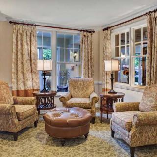 A cozy living room with armchairs and a circular coffee table, illuminated by two table lamps beside large windows with drapes.