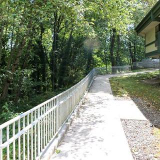 A paved path with a fence runs alongside a forested area near a building.