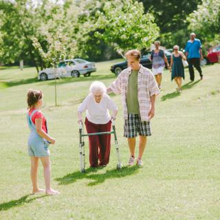 A family enjoying a sunny day in the park, with an elderly person using a walker and receiving assistance.