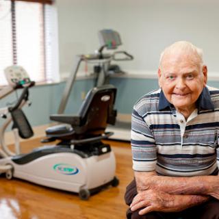 An elderly man smiles while seated in a gym, surrounded by exercise equipment designed for fitness and rehabilitation.