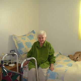 An elderly person sitting on a bed in a bedroom with a walker nearby.