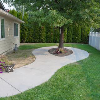 A winding concrete pathway leading through a well-groomed garden with a tree and lush green grass.