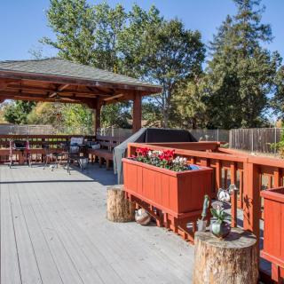 A spacious outdoor deck featuring a gazebo, grill, and seating area surrounded by planters and trees.