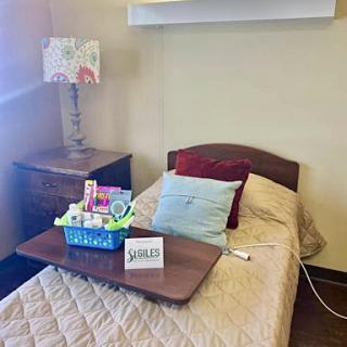 A cozy hospital room featuring a neatly made single bed with decorative pillows, a nightstand adorned with a lamp, and a tray of items.