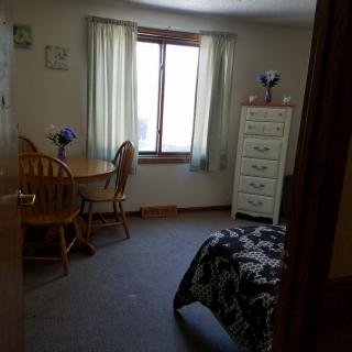 A cozy bedroom with a table and chairs, a dresser, and a neatly made bed near the window.