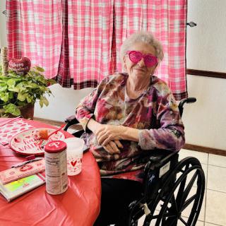 An elderly woman enjoying a festive moment at the dining table, complete with pink sunglasses and vibrant decorations.