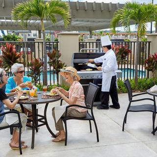 A group of seniors enjoys an outdoor meal on a patio, while a chef grills nearby.