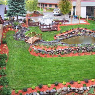 A beautifully landscaped garden featuring a pond, a wooden bridge, and well-maintained grass surrounded by colorful flower beds.