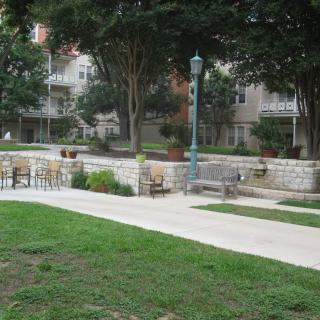 A serene park area featuring a pathway, benches, and chairs surrounded by greenery and trees.