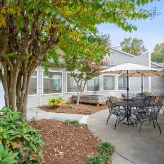 A tranquil outdoor patio area with a table, chairs, and an umbrella surrounded by lush greenery.