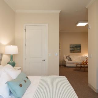 A cozy modern bedroom with a minimalist design, featuring a bed, bedside lamp, and view into a living area.