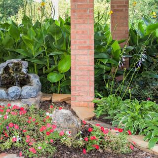 A tranquil garden scene featuring vibrant flowers, a small water fountain, and lush greenery surrounding a brick pillar.
